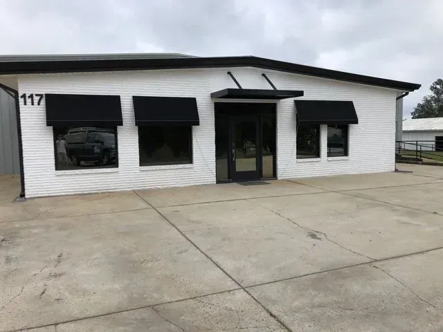Small white auto shop with black awnings and glass door, concrete lot in front, number 117 on left side.