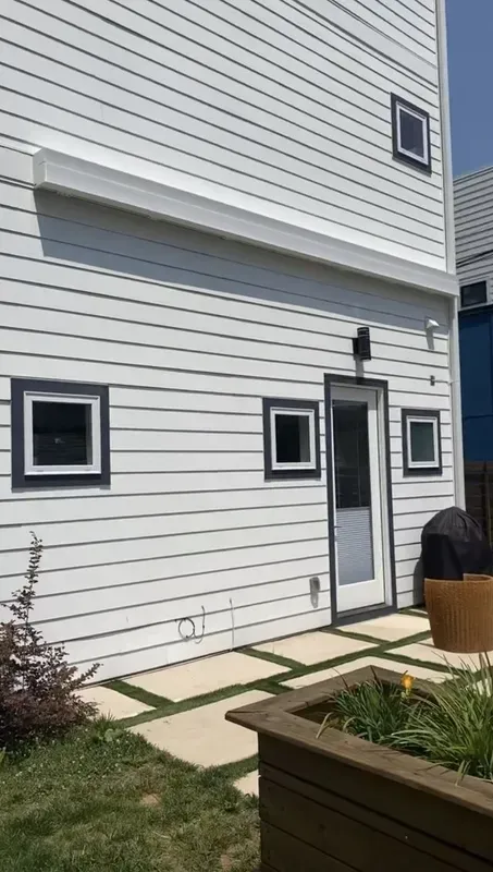 White house exterior with small square windows, side door, and a patio with planter boxes and grass.