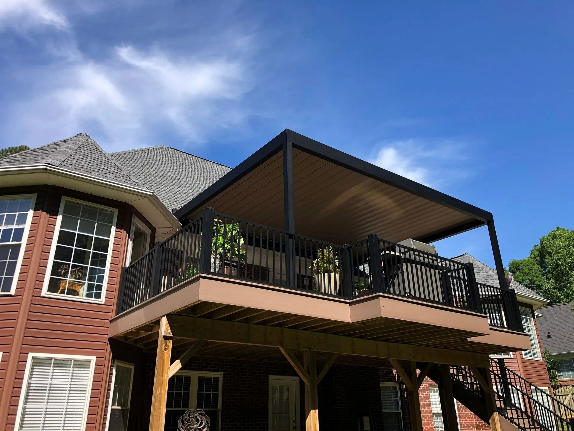 Two-story brick house with an upper deck, black railing, and gray roof under a blue sky