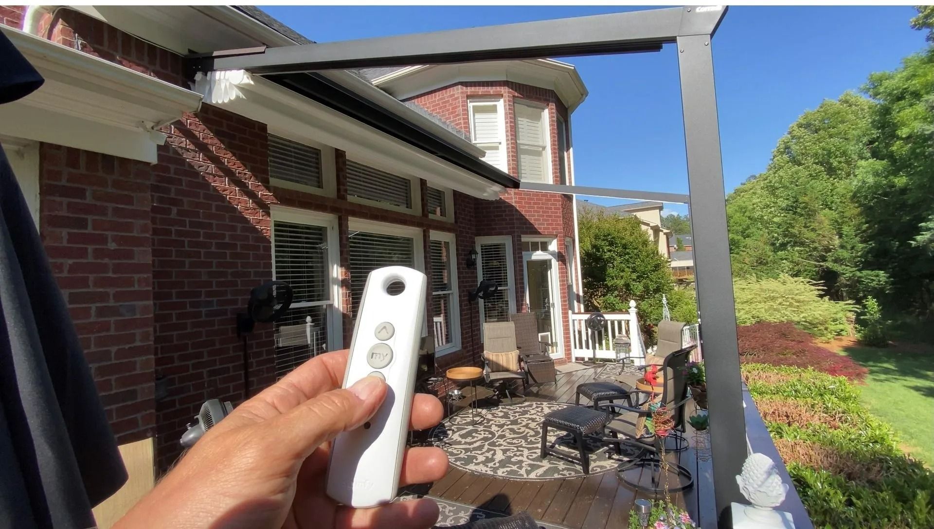Hand holding a white remote in a brick patio with pergola, outdoor chairs, and green garden