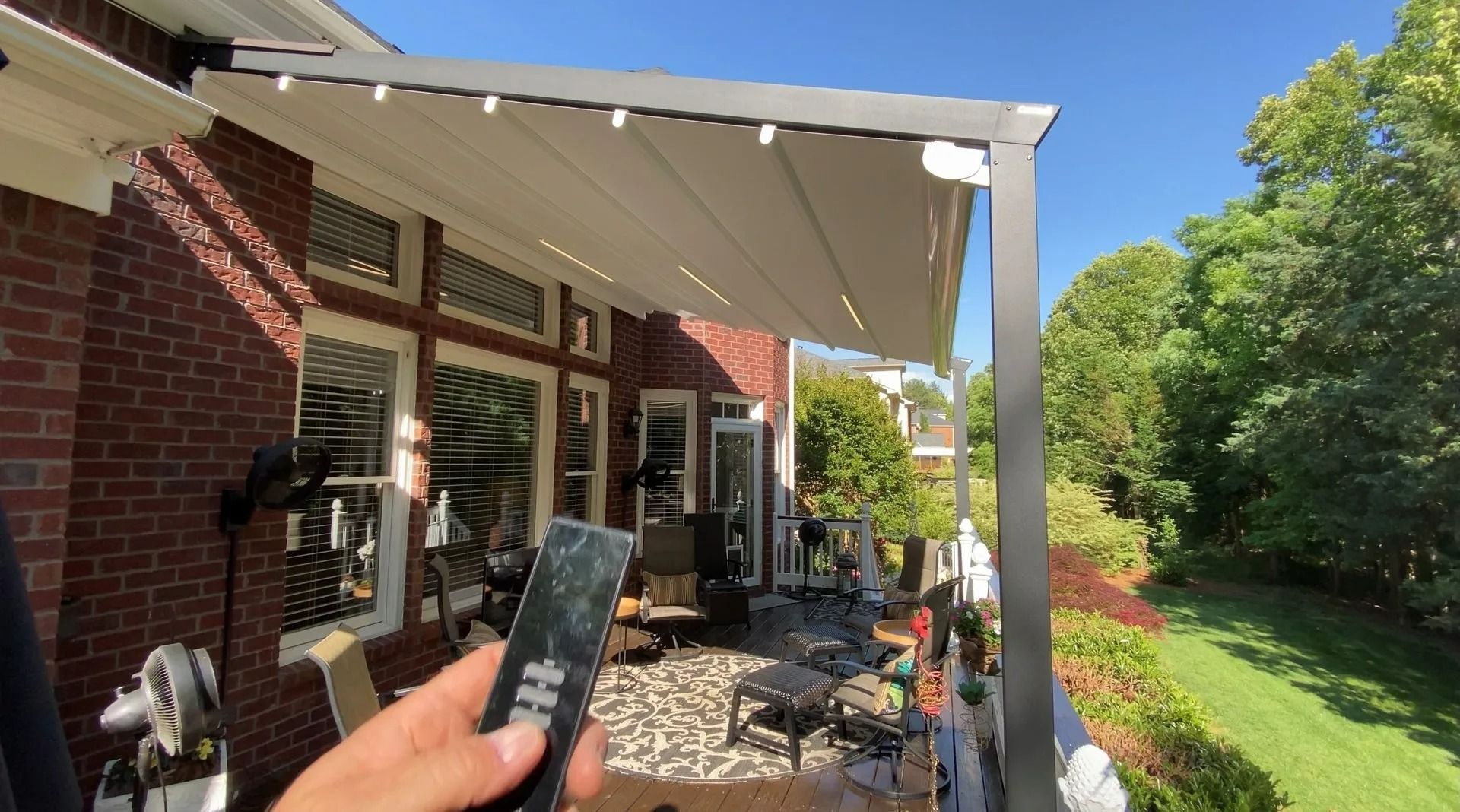 Hand holding a remote in a backyard patio with white awning, brick house, and green lawn.