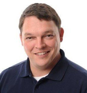 Man with short brown hair smiles, wearing a navy blue polo shirt against a white background.