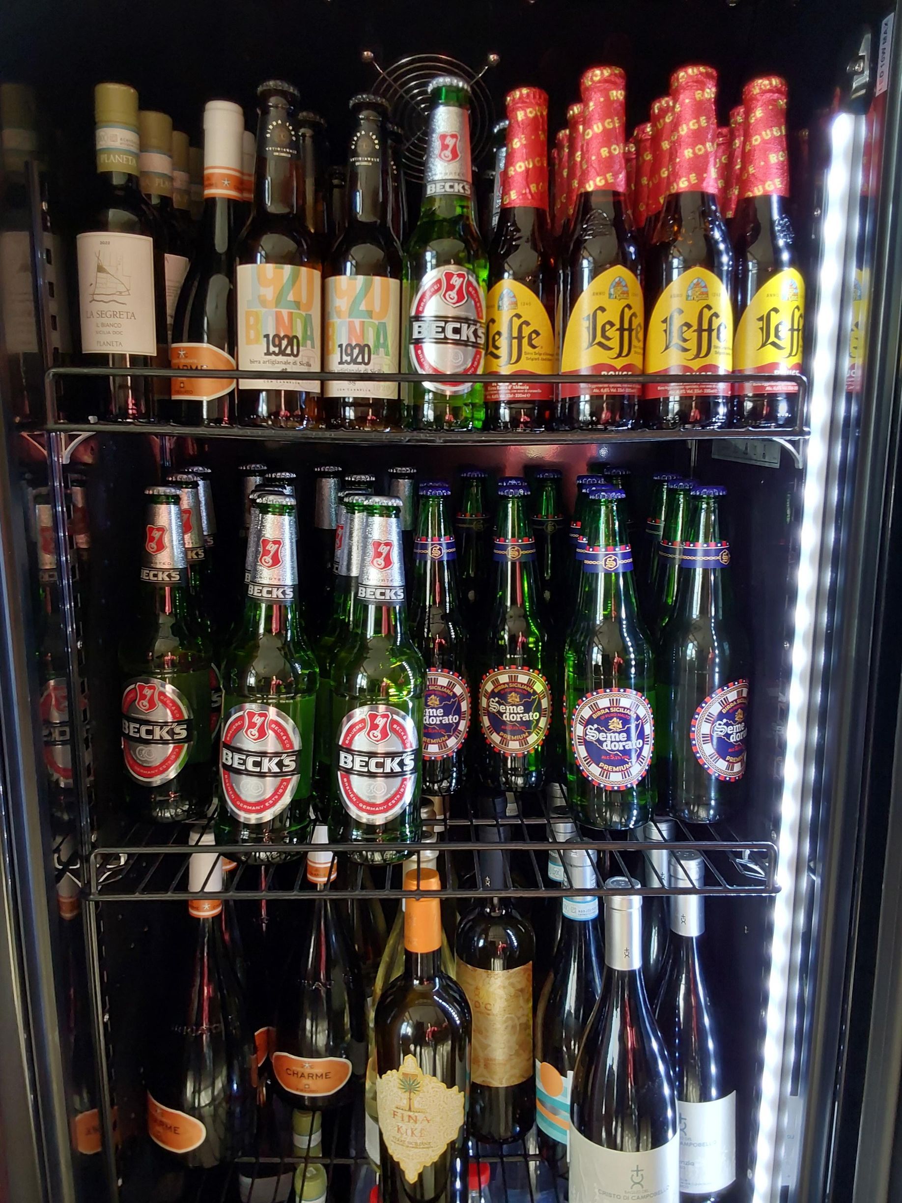 Espositore refrigerato di birre e bevande assortite in bottiglia, disposte su scaffali con etichette rosse, gialle e verdi.