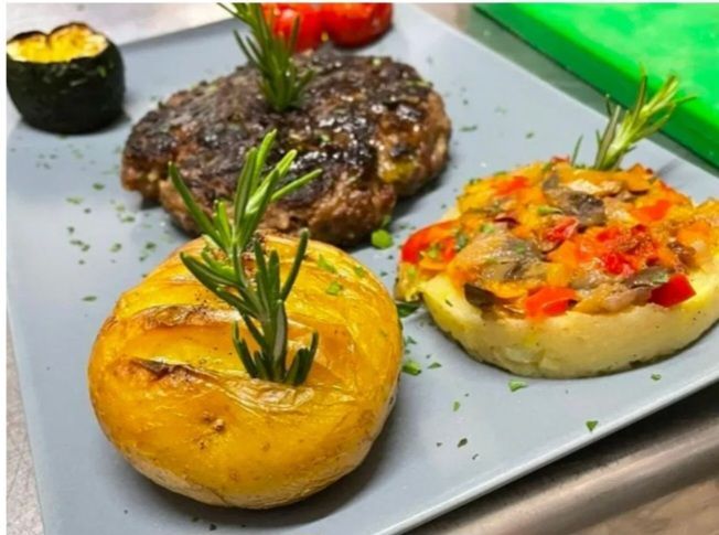 Piatto unico con due bistecche, patate arrosto e pomodoro ripieno su un piatto grigio.