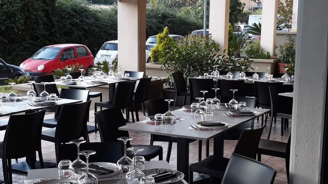 Dehor esterno del caffè con tavoli e sedie neri, apparecchiato con bicchieri, accanto a un'auto rossa e piante.