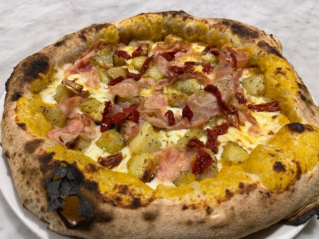 Una pizza rustica con formaggio fuso, prosciutto, salame piccante e crosta bruciacchiata su un piatto bianco.