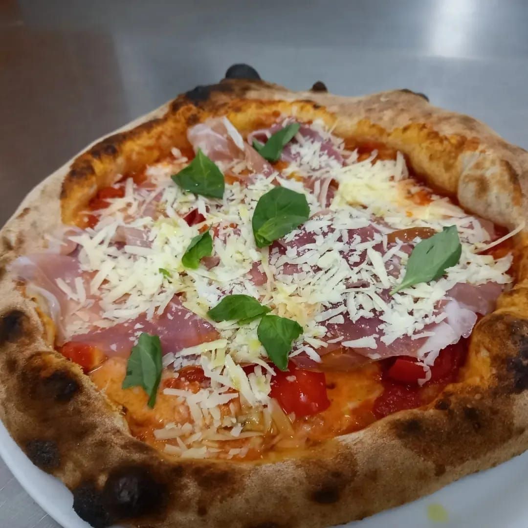 Primo piano di una pizza rustica condita con basilico, formaggio grattugiato, pomodoro e fette di prosciutto crudo.