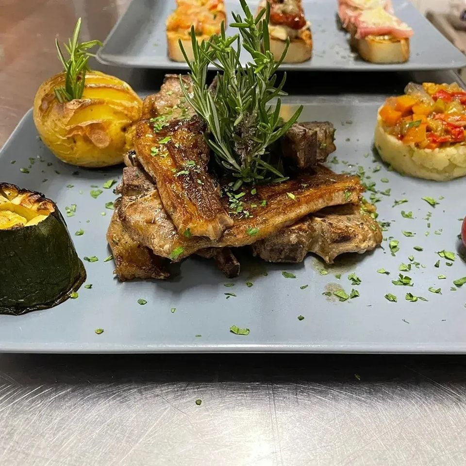 Piatto gourmet impiattato con carne grigliata, rosmarino, verdure e guarnizioni colorate su un vassoio grigio.