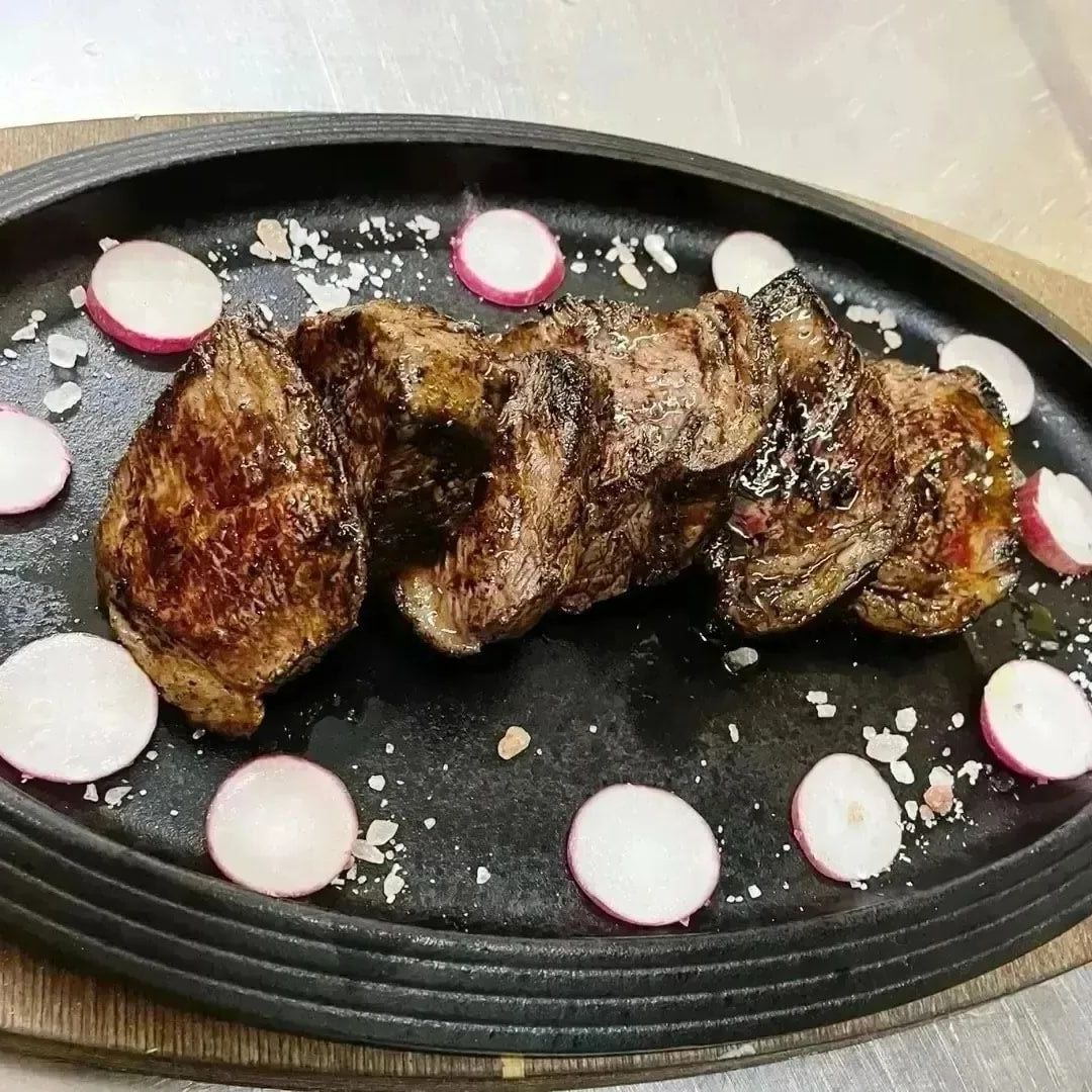 Bistecca alla griglia servita su un piatto ovale nero, guarnita con ravanelli a fette e sale grosso.