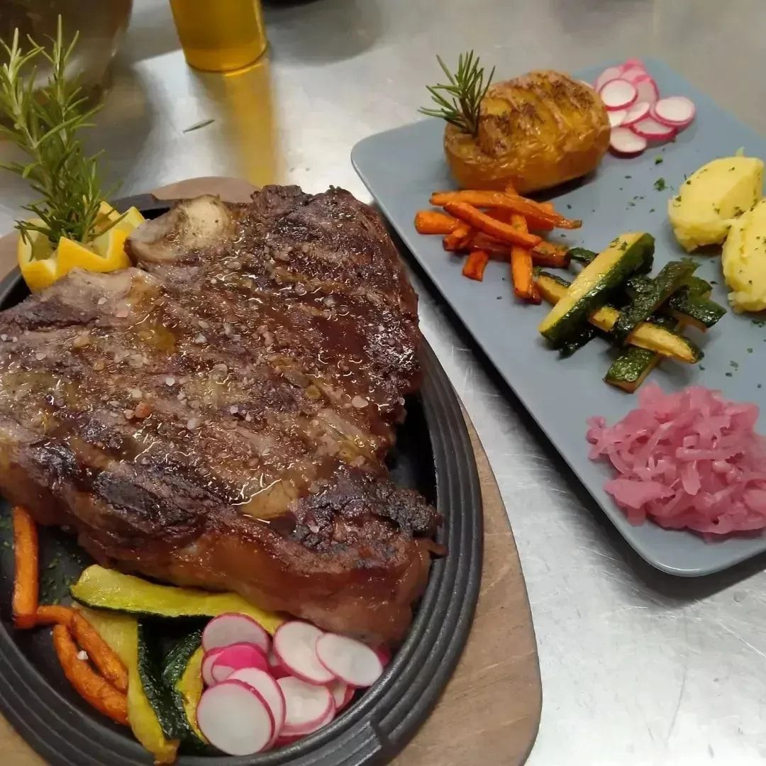 Bistecca alla griglia con verdure arrosto e patate su vassoi impiattati in un ambiente di cucina.