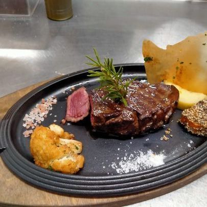 Bistecca impiattata con guarnizione di rosmarino, carne affettata e contorni su un piatto nero in un ambiente di cucina.