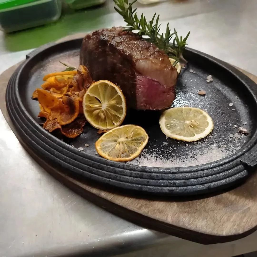 Bistecca scottata su un piatto nero con fette di limone, erbe aromatiche e guarnizione croccante su un bancone in acciaio inox.