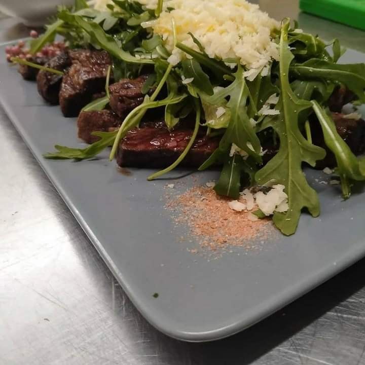 Fettine di bistecca grigliate con rucola e formaggio grattugiato su un piatto grigio, con condimento a parte.