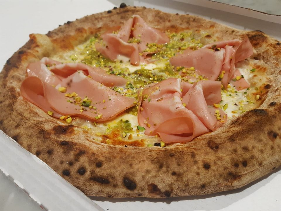 Pizza cotta nel forno a legna con prosciutto, formaggio ed erbe aromatiche, servita su un piatto bianco.