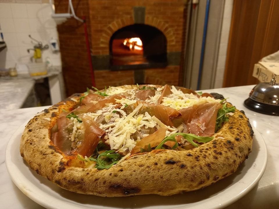 Pizza cotta nel forno a legna, condita con prosciutto e rucola, servita su un piatto accanto al forno fumante.