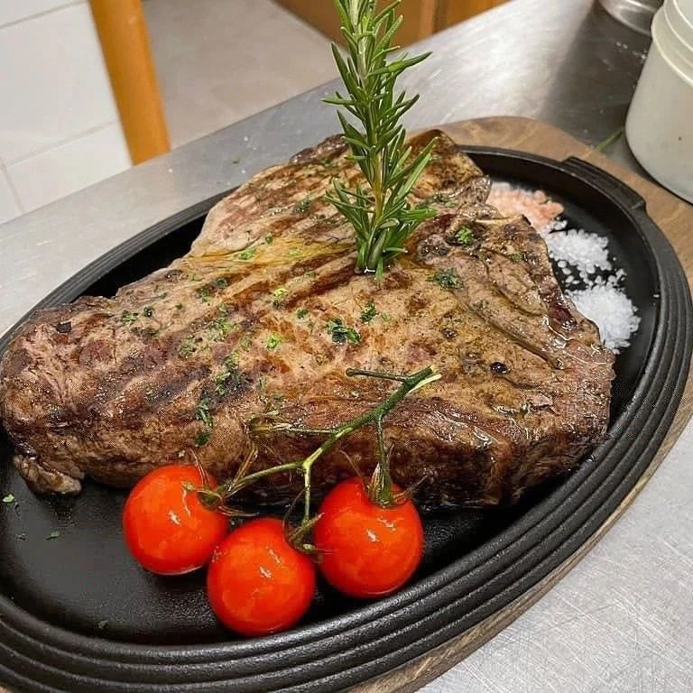 Bistecca alla griglia su un piatto nero con rametto di rosmarino e pomodorini su un bancone in acciaio inox.