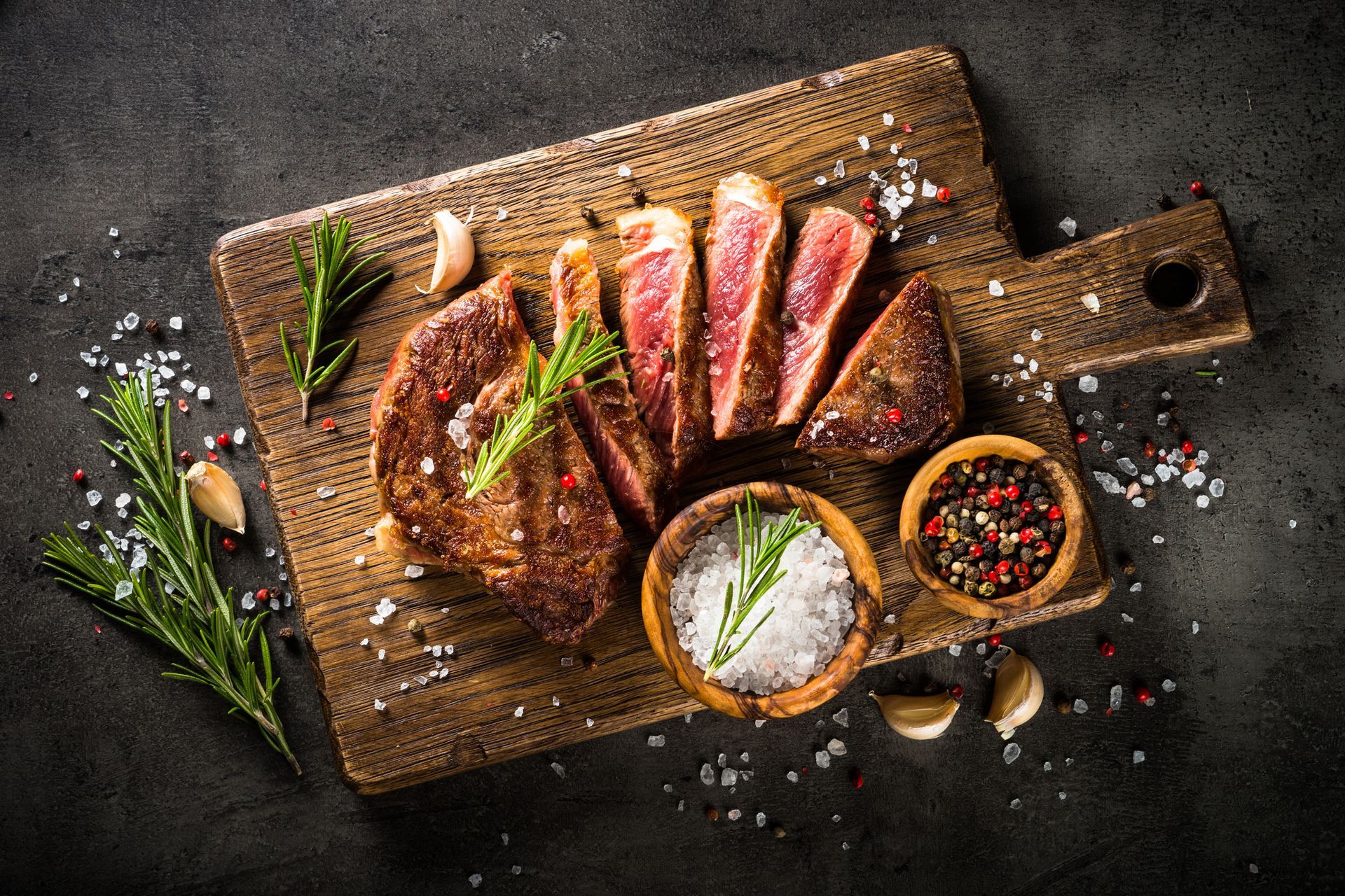 Fettine di bistecca su un tagliere di legno con rosmarino, burro, grani di pepe ed erbe aromatiche su un piano di tavolo scuro