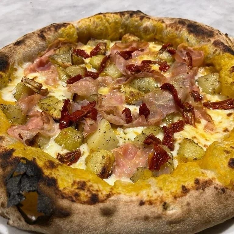 Primo piano di una pizza gourmet con formaggio fuso, prosciutto, patate e peperoni rossi su una crosta bruciacchiata.