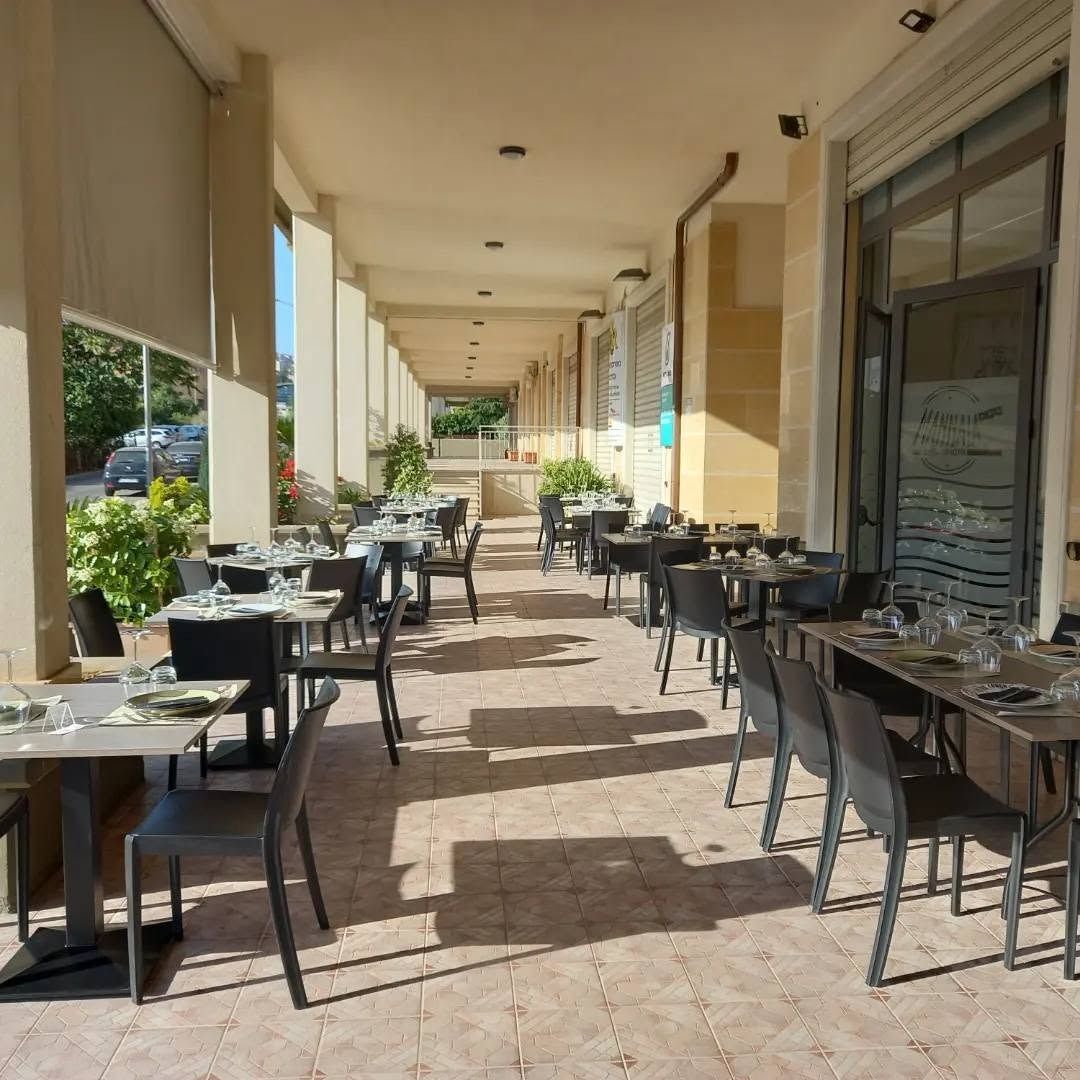 Terrazza esterna del caffè con tavoli e sedie neri sotto un passaggio coperto, soleggiata e vuota.
