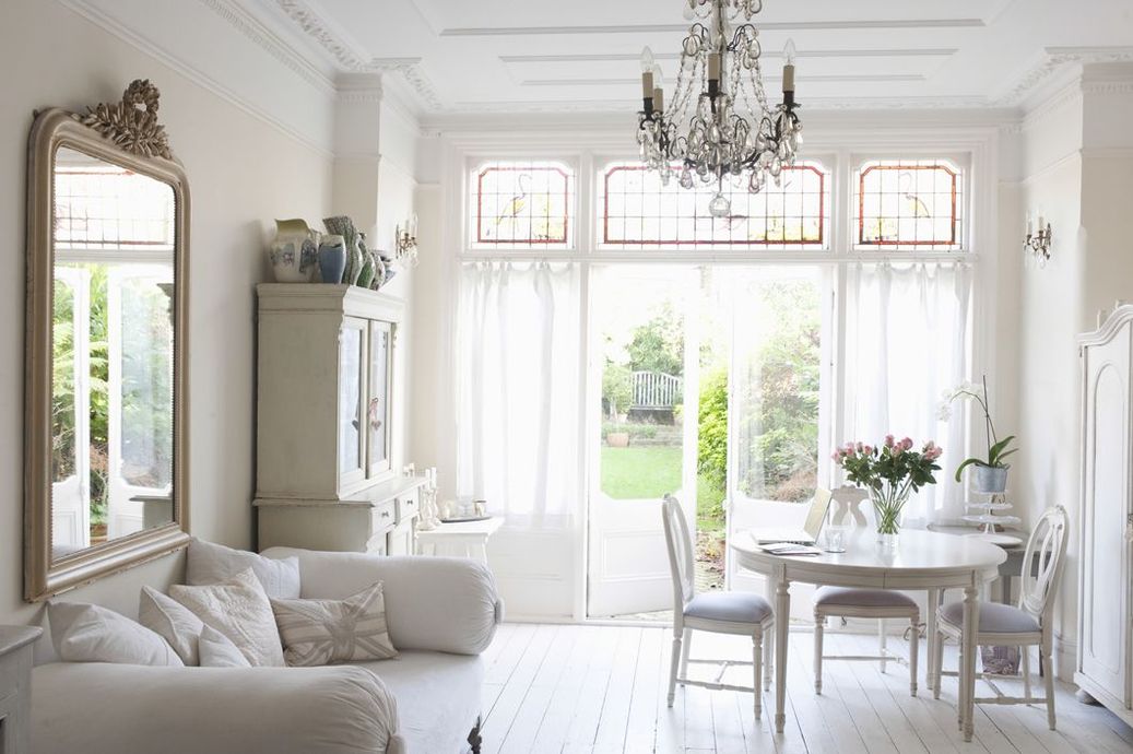 Bright white elegant living room with chandelier, sofa, dining table, and tall windows opening to a garden.
