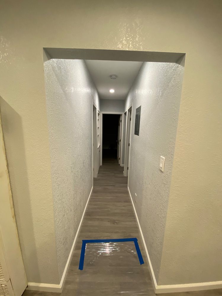 Narrow apartment hallway with white walls, gray carpet, and blue tape marking the floor in the foreground