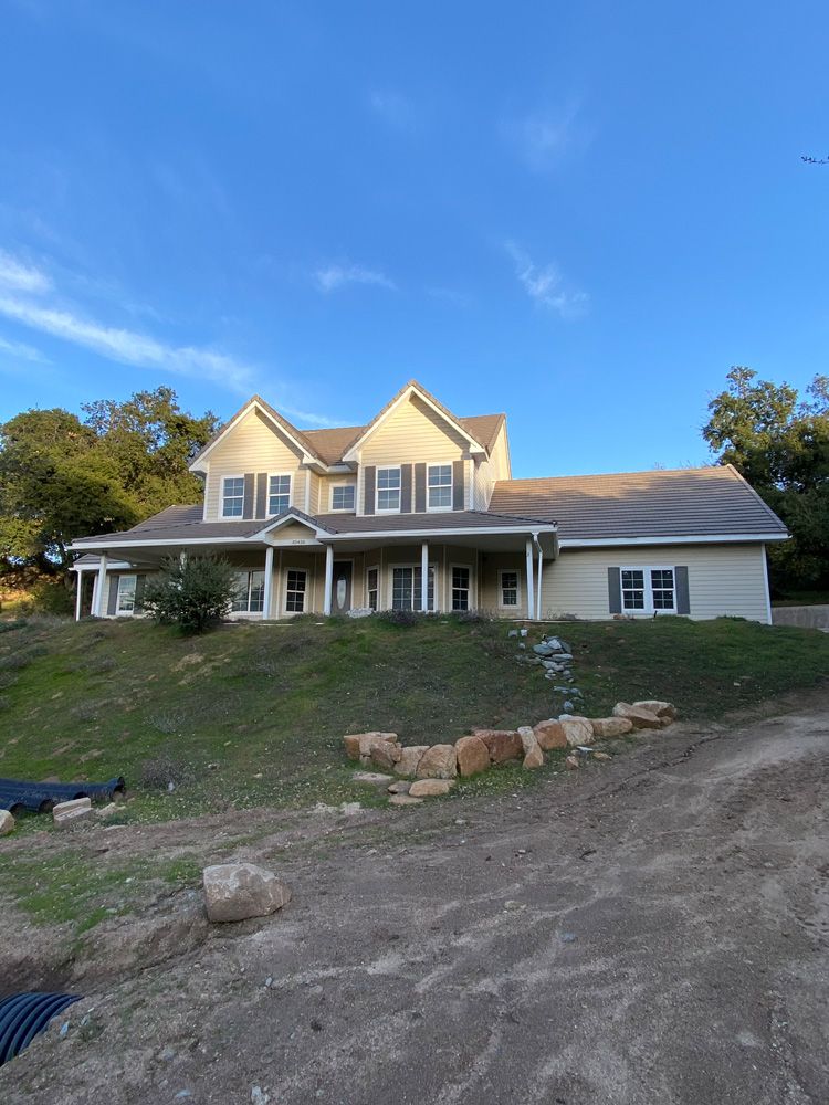 Two-story white house with porch on a sloped dirt lot under a blue sky