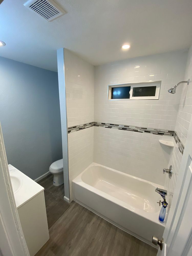 Bathroom with white tub, tiled shower wall, small window, and wood-look floor.