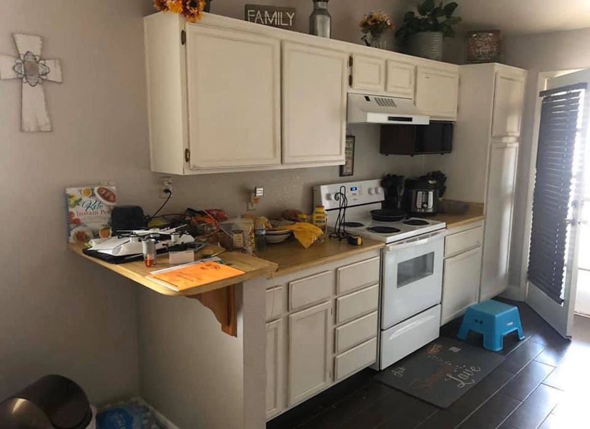 Small kitchen with white cabinets, stove, countertop clutter, and a blue step stool by the window blinds.
