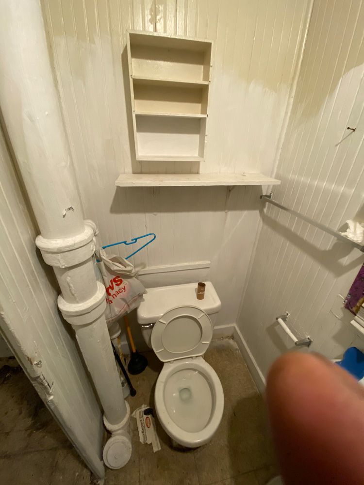Small bathroom with toilet, shelves, pipes, and sink; a finger partially covers the right side.