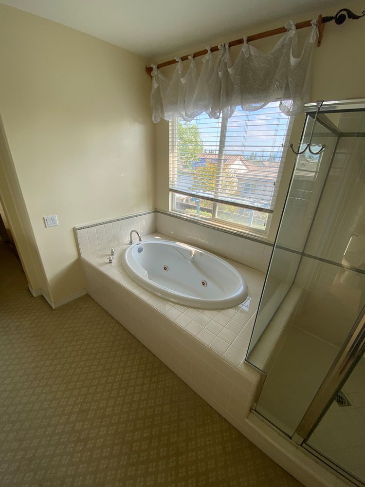 Bathroom with built-in jetted tub, glass shower, and window with blinds and sheer curtains.