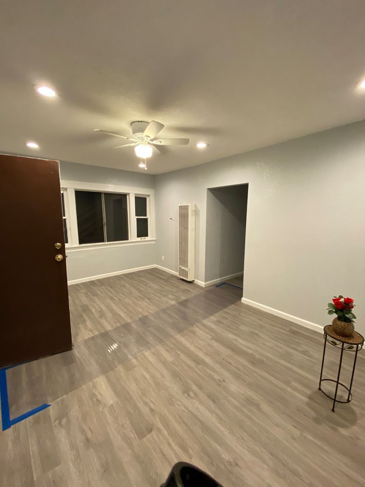Empty living room with gray wood floors, white walls, ceiling fan, window, and small plant stand.