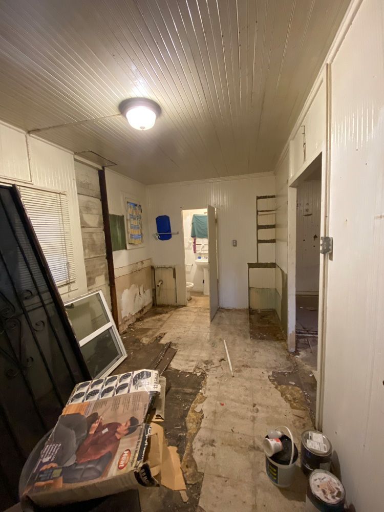 Narrow, cluttered hallway with peeling walls, damaged floor, and a chair covered by a patterned cloth.