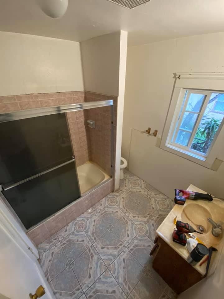 Small bathroom with glass shower enclosure, patterned tile floor, window, and vanity clutter.