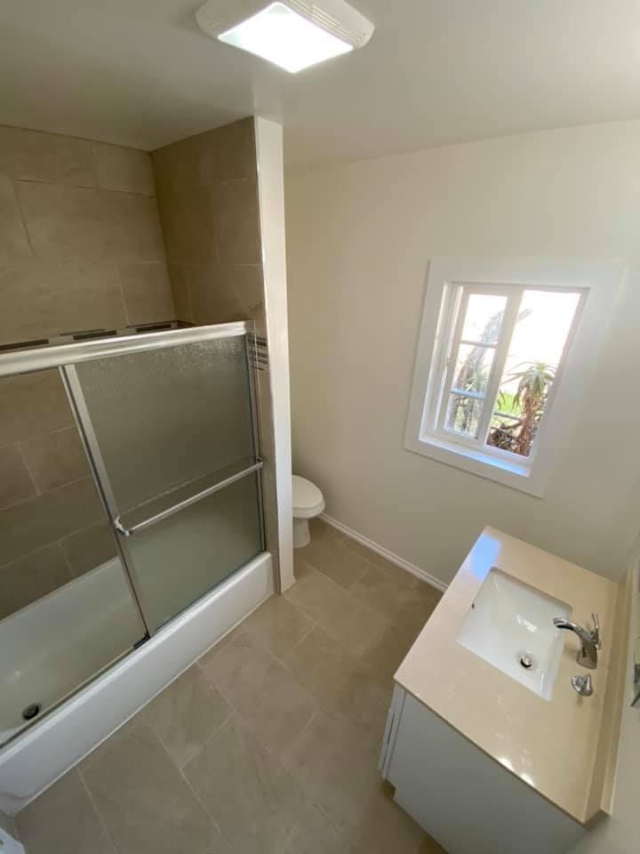 Small bathroom with shower, toilet, sink cabinet, beige tiles, and a window letting in daylight.
