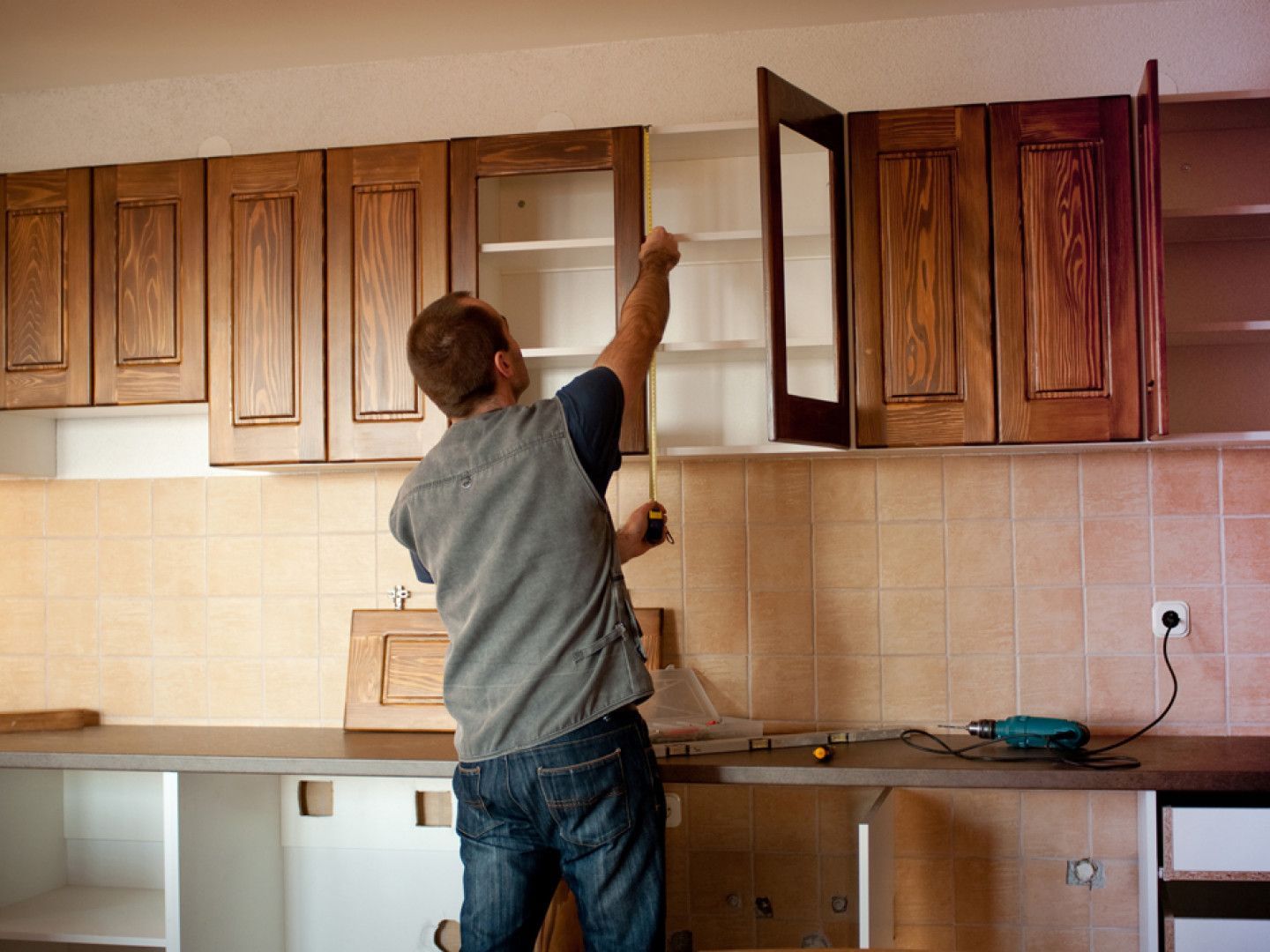 Person reaching into an open kitchen cabinet during installation, with unfinished countertop and tiled backsplash.