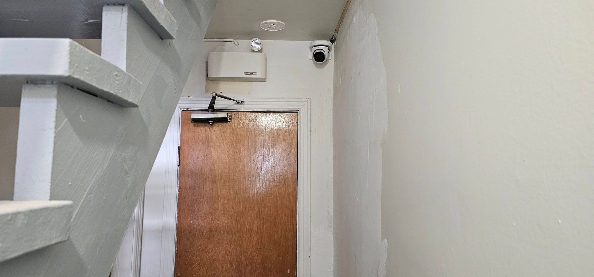 Une porte marron avec un ferme-porte automatique se trouve sous un escalier, à côté d'un mur où se trouvent un boîtier blanc et une petite caméra de sécurité.