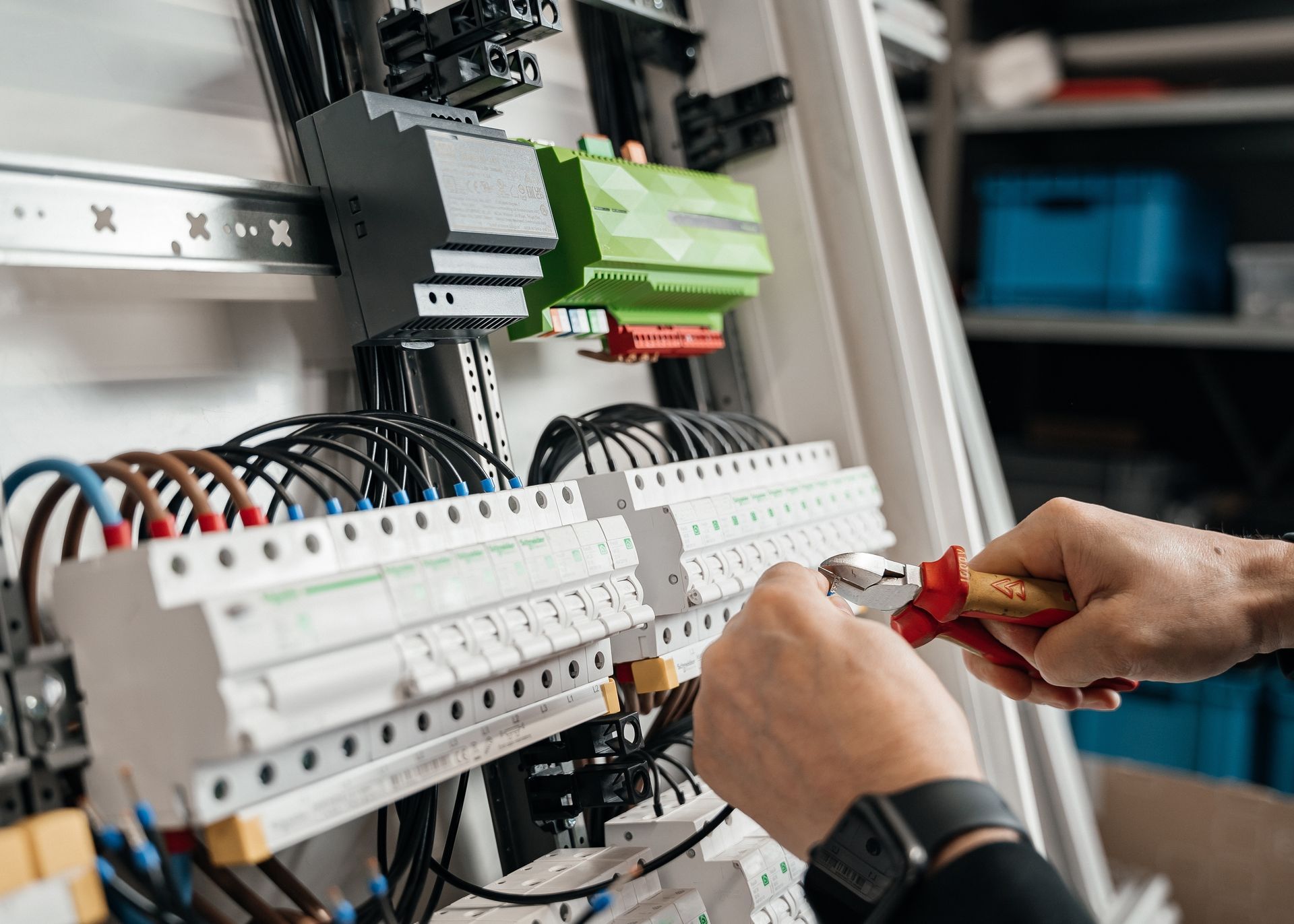 Des mains manipulent des pinces coupantes à poignée rouge pour travailler sur des composants électriques dans un tableau de disjoncteurs.