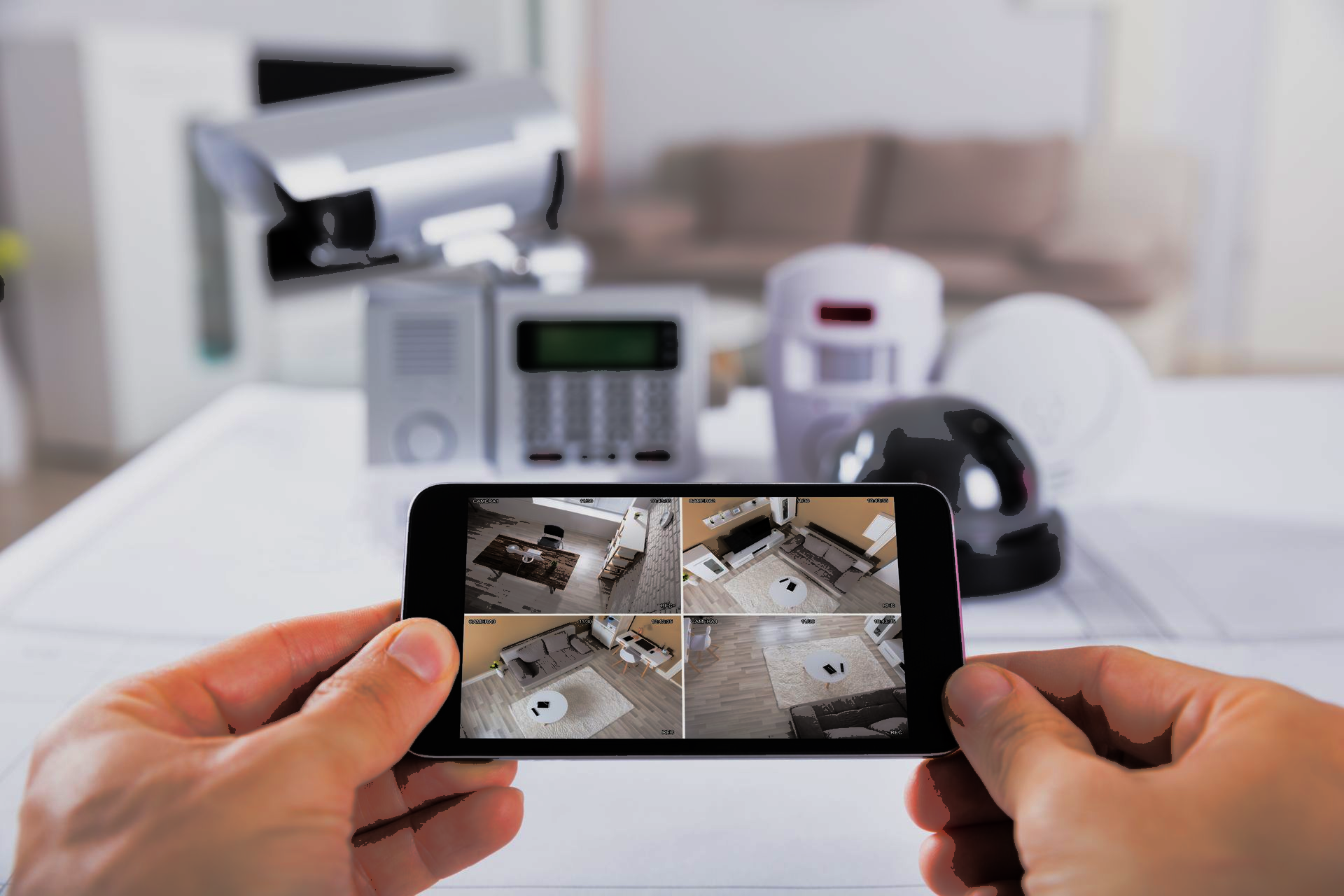 Des mains tenant un smartphone affichant les images en direct d'une caméra de sécurité, avec du matériel de sécurité domestique flou en arrière-plan.