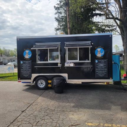 Black food truck with serving windows and menu signs parked on a paved lot outdoors