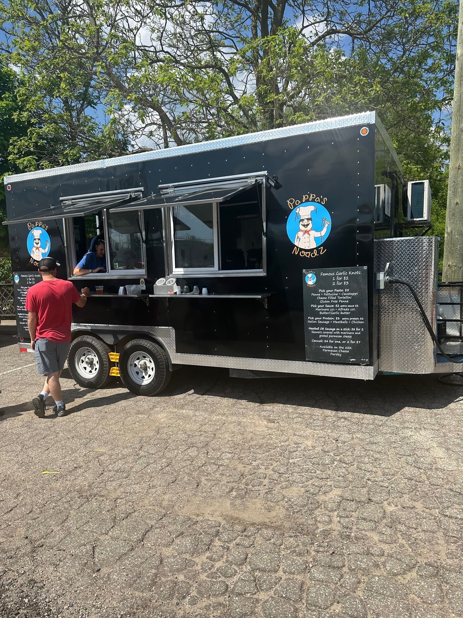 Black food trailer with awning and menu board at outdoor market, person in red shirt standing nearby