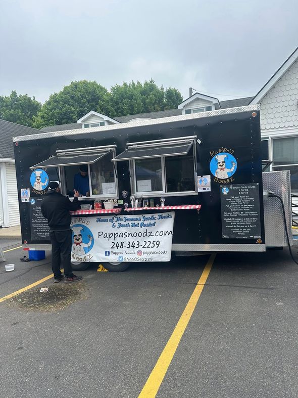 Black food truck with service window and menu signs parked in a lot, person standing beside it