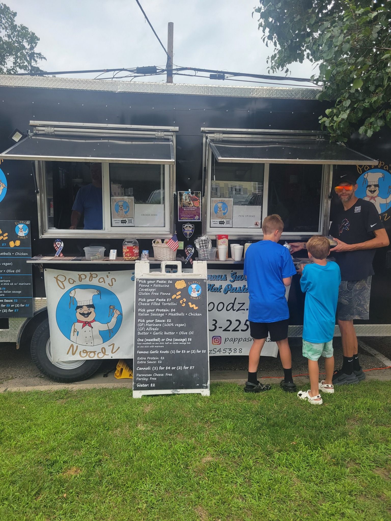 Food truck window serving customers outdoors, with two children ordering at a chalkboard menu.