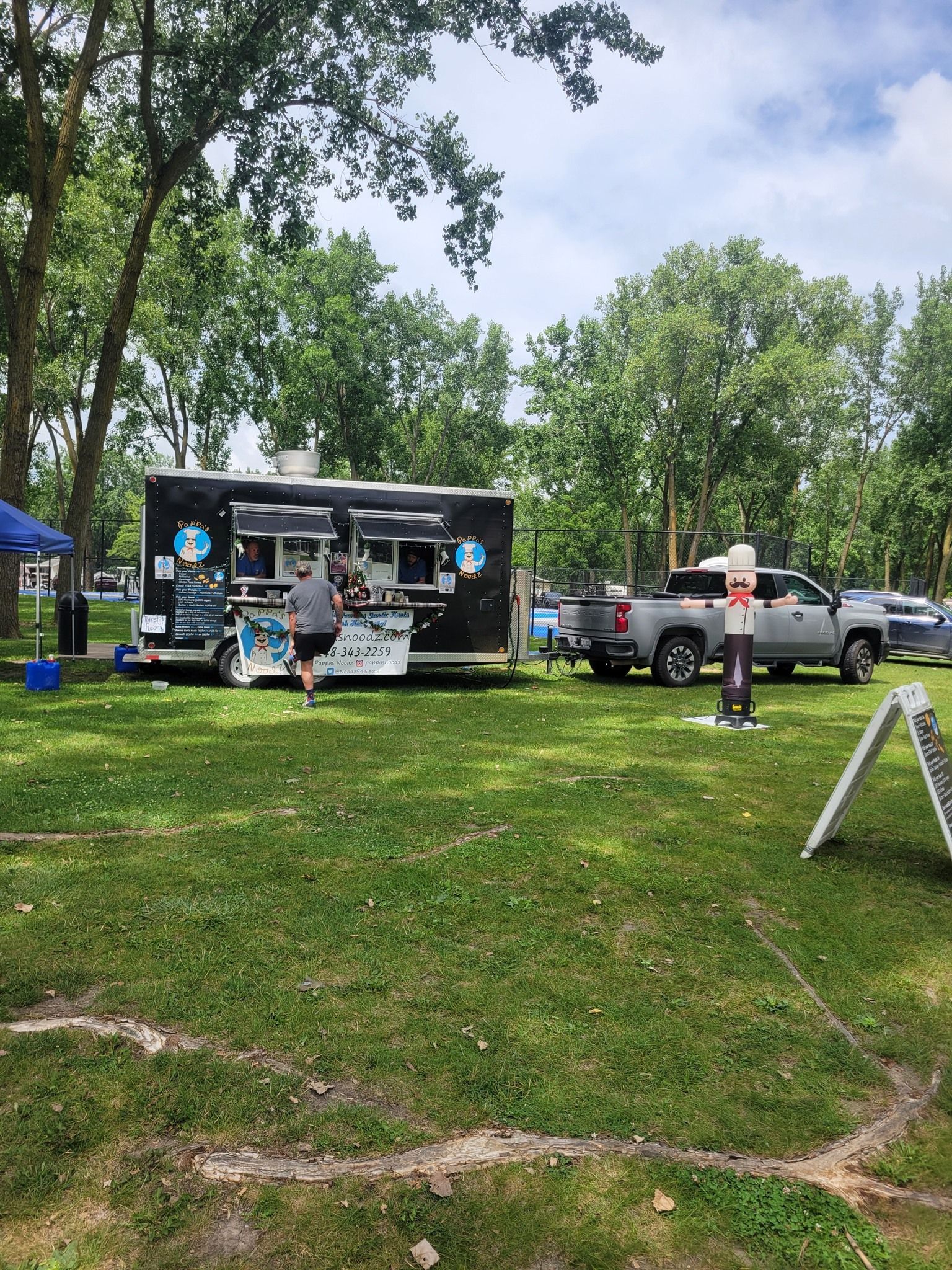 Outdoor park event with a food truck, people gathered, and a pickup truck nearby under trees.