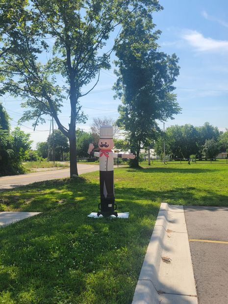 Person standing on grass beside a road, holding a sign in a sunny park with trees.