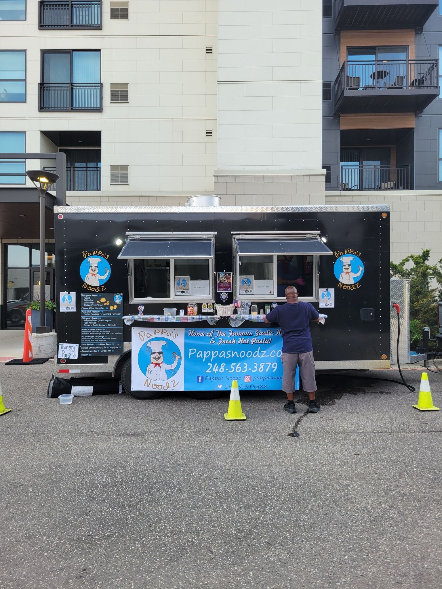 Food truck with blue signage parked outside apartments, with a person standing at the counter and cones in front.