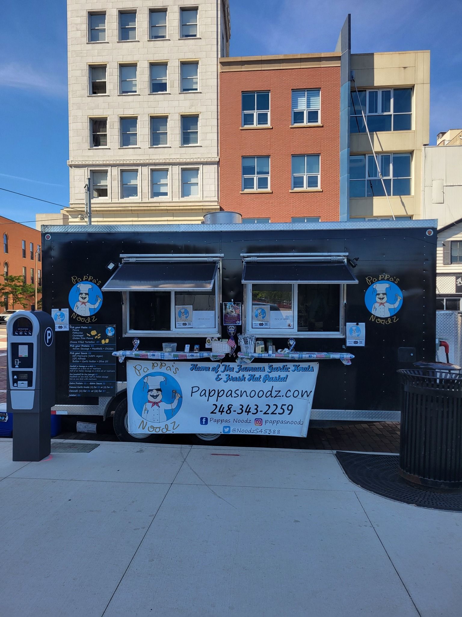Food truck with blue awning and banner parked on a city sidewalk in front of buildings.