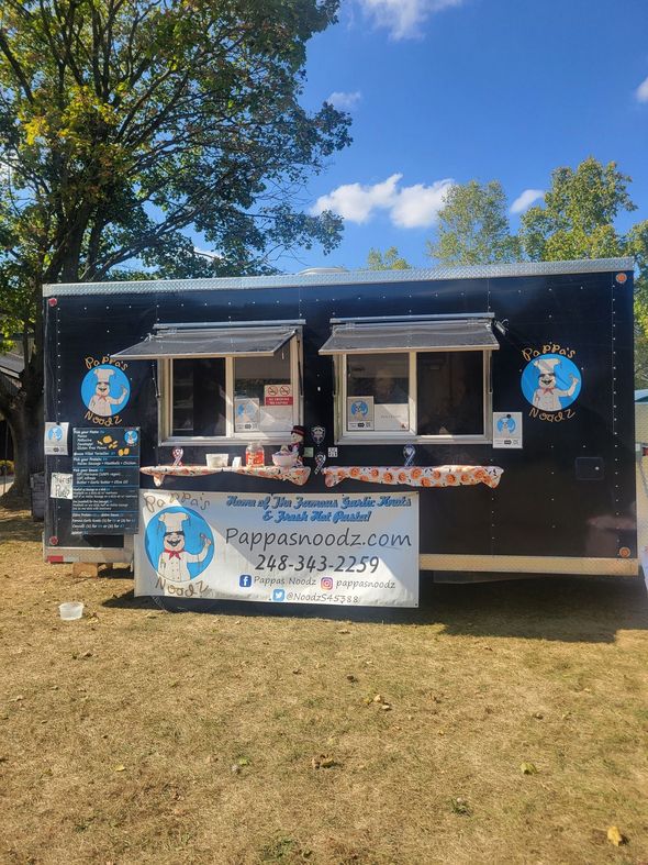 Black food trailer with service windows and blue snowman logos on a grassy field under trees and blue sky