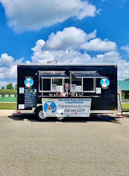 Black food trailer with serving windows and banner parked on a gravel lot under a blue sky.