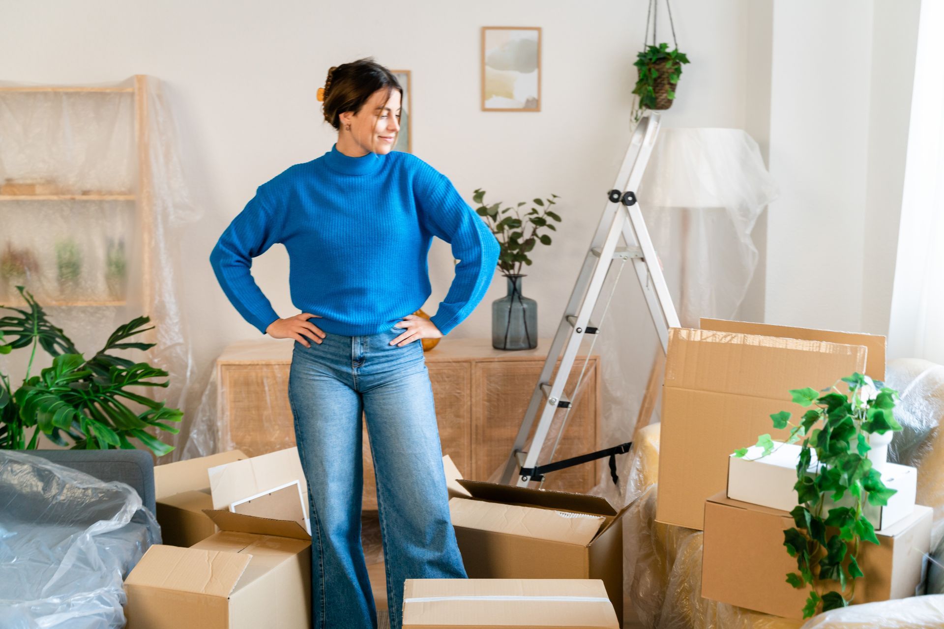 A woman in a blue sweater is standing in a living room surrounded by cardboard boxes.