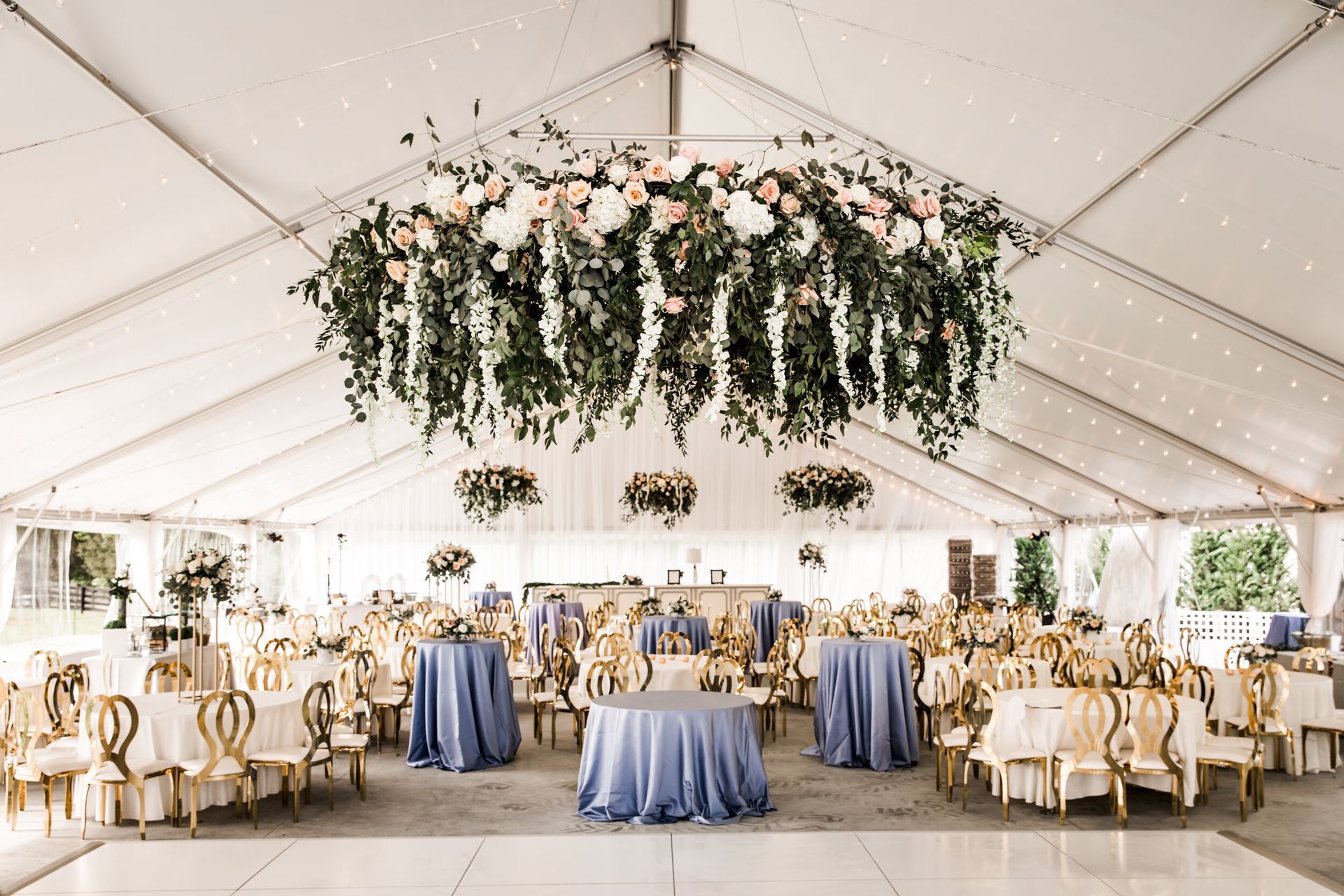 Wedding reception in a tent. Tables with blue cloths, gold chairs, and hanging floral arrangements.